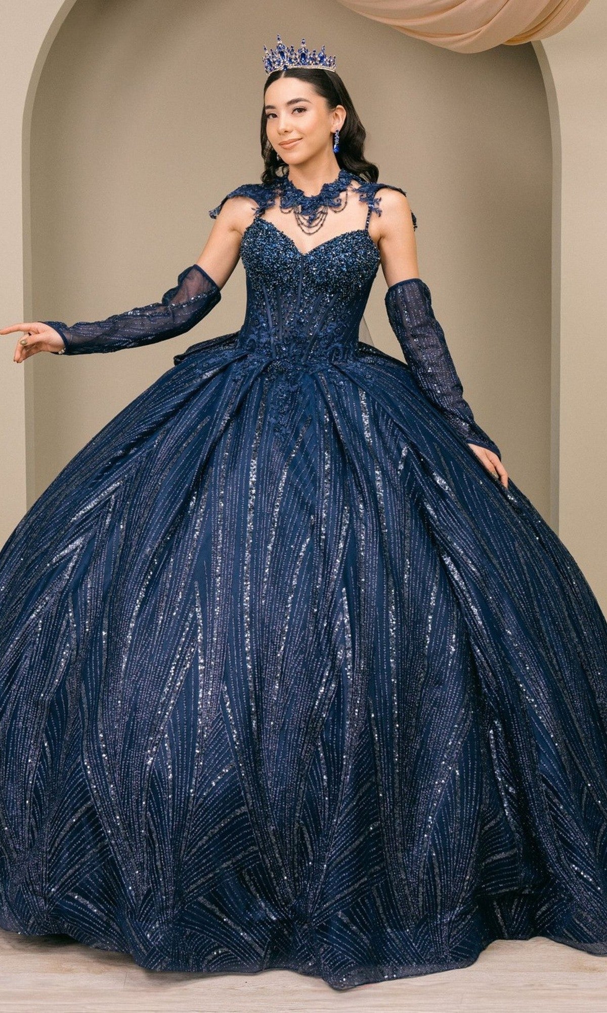 A woman wearing the Quinceanera Dress 8510JQ by Cinderella Couture in navy, adorned with sequin embellishments, along with matching gloves, necklace, and tiara, stands indoors against a neutral background.